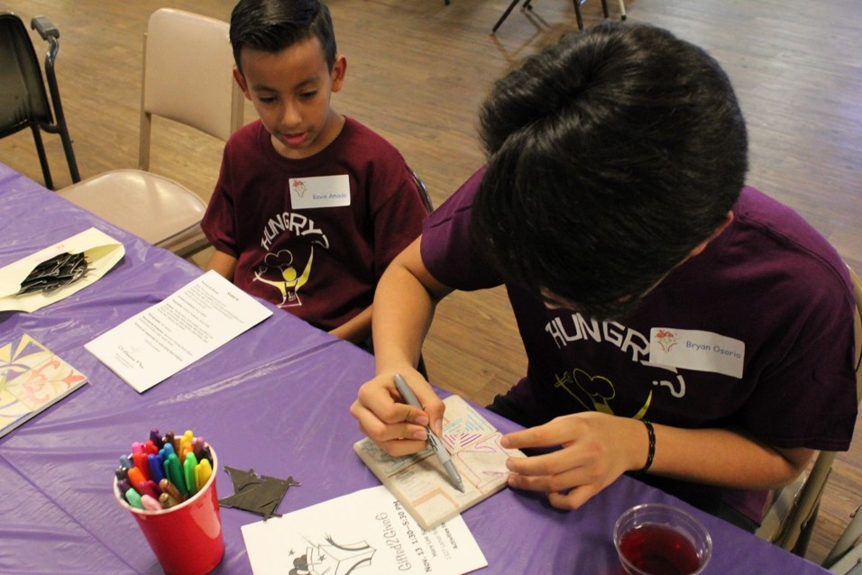 Two young artisans in purple Spacious Place t-shirts seated and creating ceramic tile art.