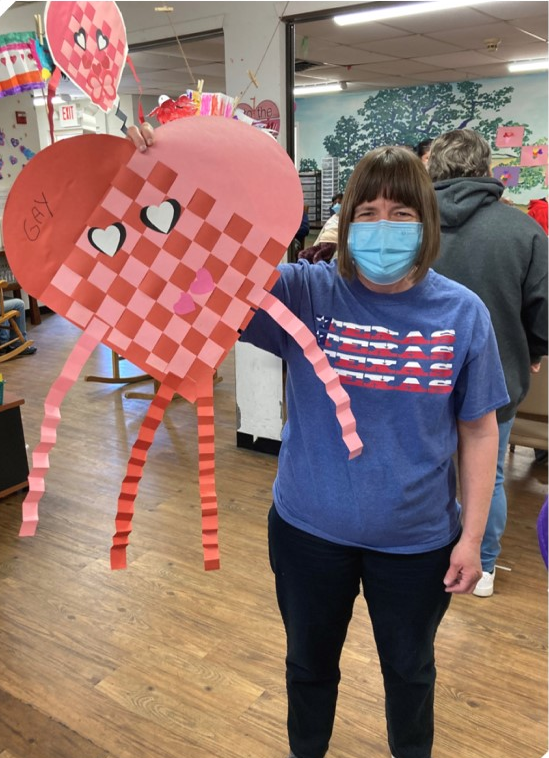 Participant masked and holding a giant red and pink Swedish Heart person with face, arms, and legs. created these for Valentine's Day.