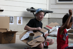 Camper in a fedora playing a Picasso-style cardboard and string while another camper measures more string.