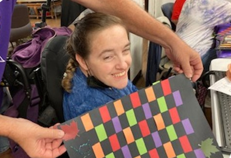 Participant, seated in a wheelchair, smiles as a guide holds up the paper weaving they created together. The frame paper is black and the paper strips are in fall colors.