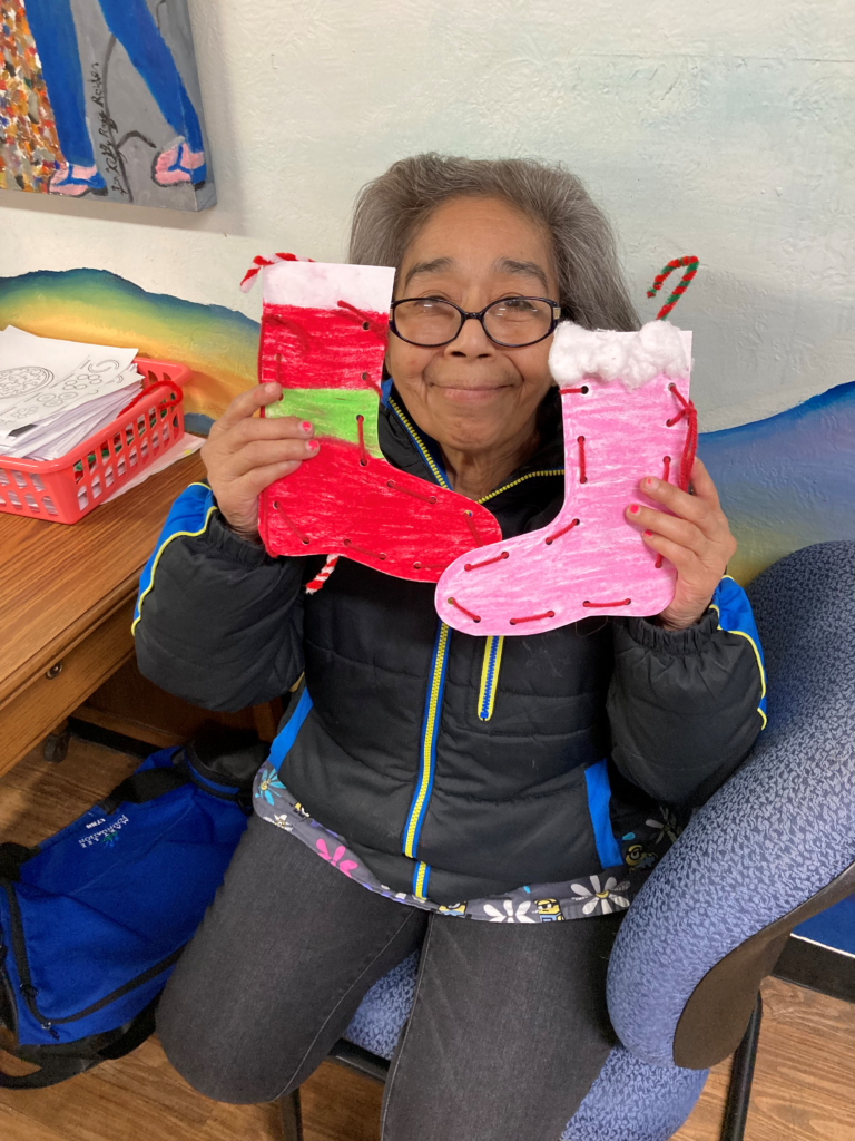 Seated and smiling participant holds two paper stockings she made, decorated and laced. Each contains a chenille-stem candy cane.