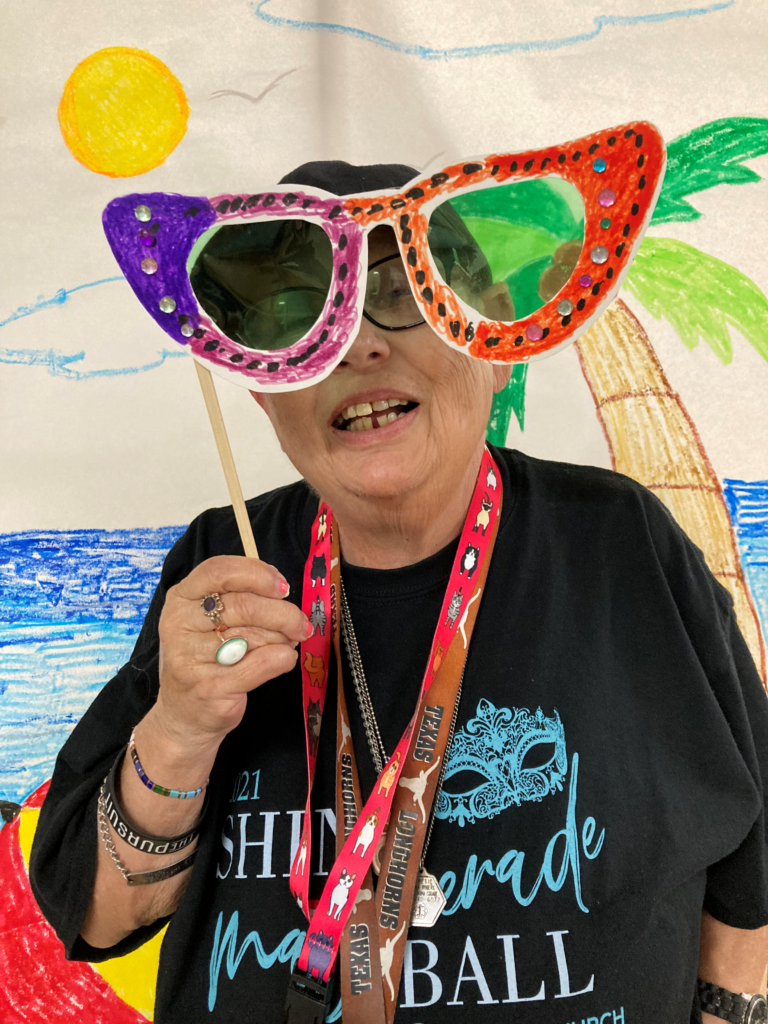 Participant modeling the super-sized pair of sunglasses she created from poster board, cellophane, markers, gems, and glitter glue. Participant poses in front of a beach scene.