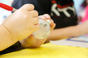 Hands are decorating a white frosted christmas ornament with holly using a red marker.